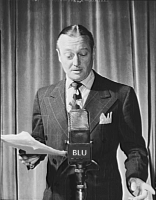 Edmund Lowe, attore americano, in piedi davanti a un microfono radiofonico (etichettato per la rete Blue Network della NBC), con in mano un copione, 1942
