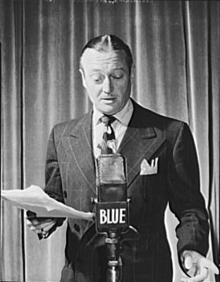 Edmund Lowe, American actor, standing at radio microphone (labeled for (NBC) Blue Network), holding script, 1942